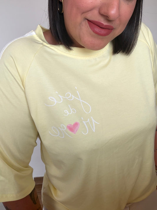 Primer plano de camiseta amarilla pastel con mensaje joie de vivre, corazón y franjas blancas