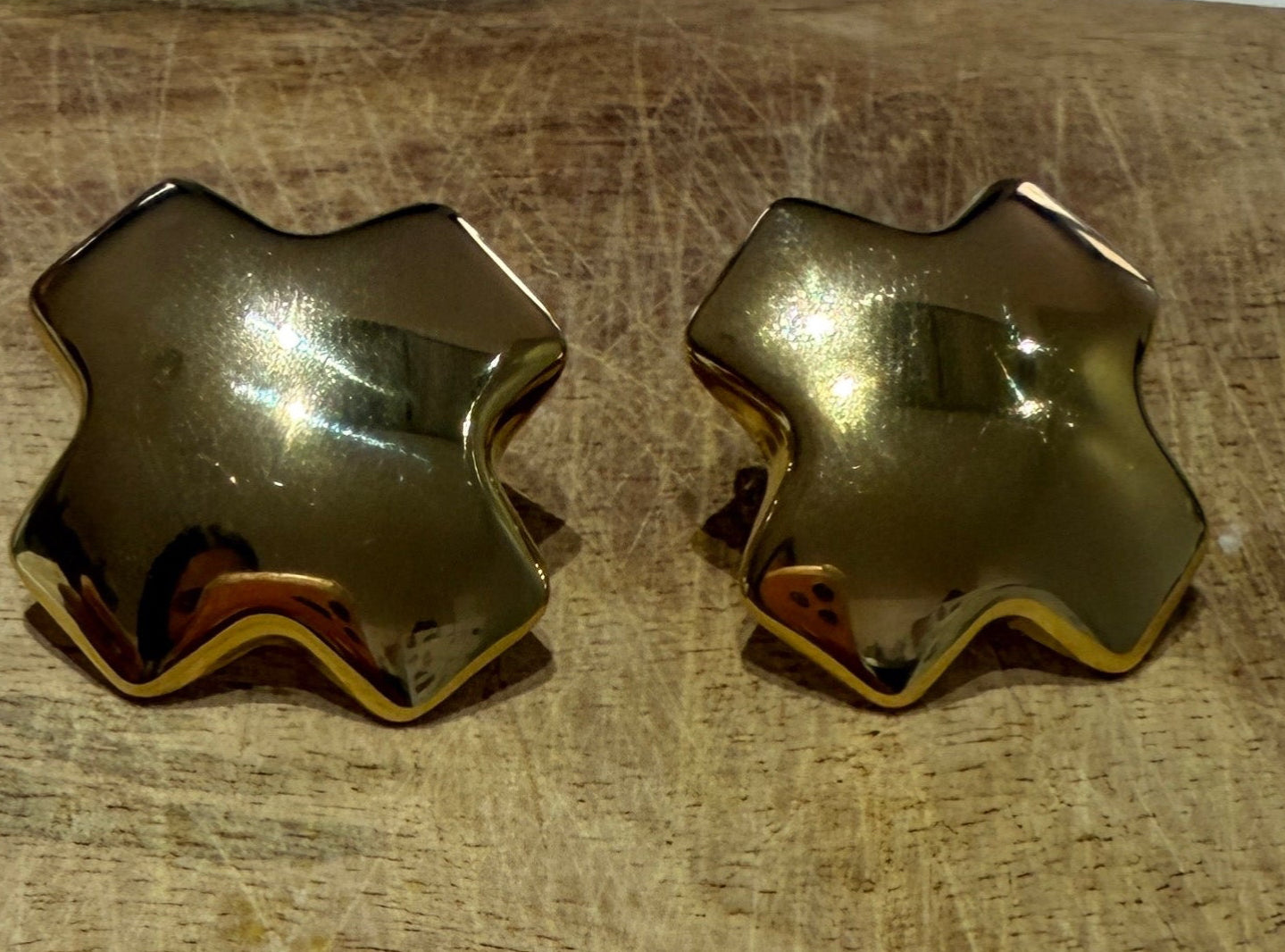 Detalle de pendientes dorados geométricos con forma de cruz y acabado brillo espejo sobre madera